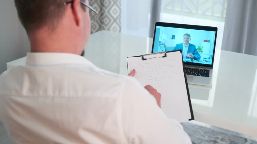 Rear view. An HR specialist conducts an online interview at his desk, with the candidate on the laptop screen, asking questions, taking notes on a clipboard, and recording key interview details.