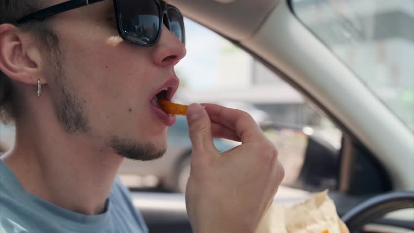 Side view. The young man in sunglasses holds a kraft paper with fries, dipping one into ketchup before bringing it to his mouth, enjoying the snack in his car on a sunny day.