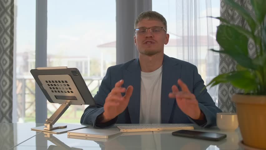 A young teacher in glasses leads an online webinar, smiling as he explains the material. He switches slides on his tablet, displaying examples and illustrations to deepen understanding of the topic.