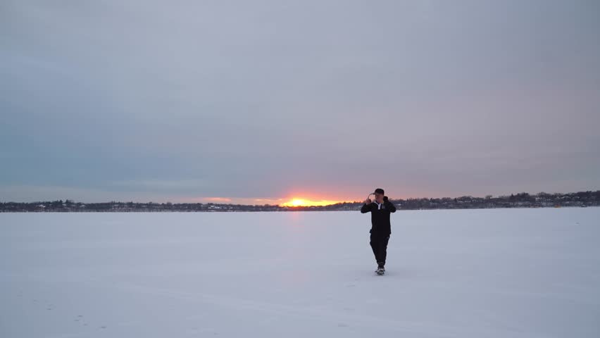 A man playing with his pug dog on the frozen lake in Minnesota, with the golden sunset in the background