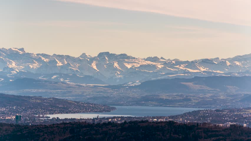 A stunning panoramic view of snow-capped peaks surrounding Zurich, Switzerland. Timelapse of the golden sunlight casting warm hues over the snow covered mountains. Boats move across Lake Zurich.