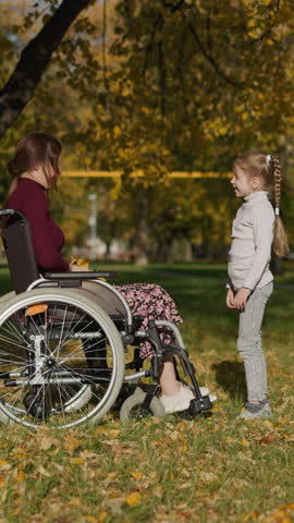 Young mother with legs injury throws bunch of dry leaves up above daughter. Preschooler girl raises hands rejoicing at game in autumn park side view