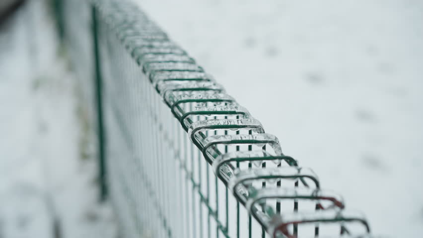 Close-up of green metal fence coated with ice, showcasing icicles hanging from the wires against a blurred snowy park background, emphasizing frozen textures and the stark coldness of winter