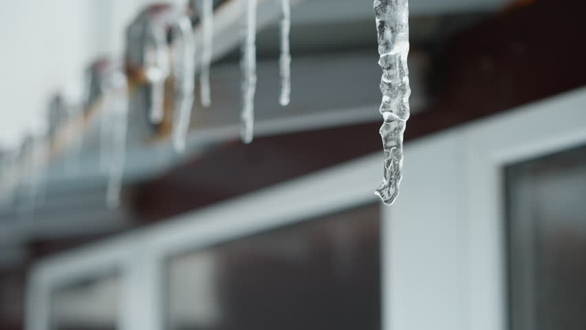 Close-up of house metal roof edge with long icicles melting and dripping water droplets, highlighting intricate frozen structures against blurred background