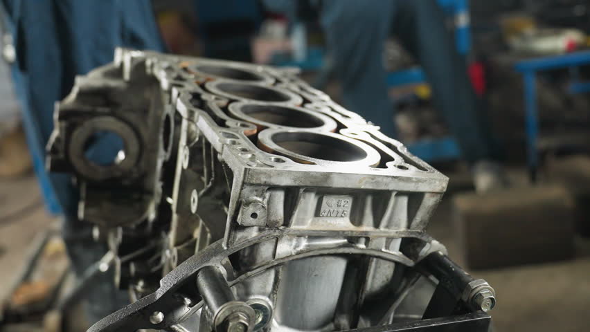 Close-up of car engine with metallic details, bolts, and cylinders, placed on a stand, people in blue uniforms visible in background working in an automotive workshop filled with mechanical tools