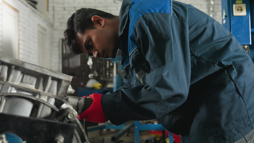 Mechanic in red gloves uses pneumatic tool on engine block in industrial workshop, performing precise mechanical repair, focused worker in blue uniform adjusts engine components