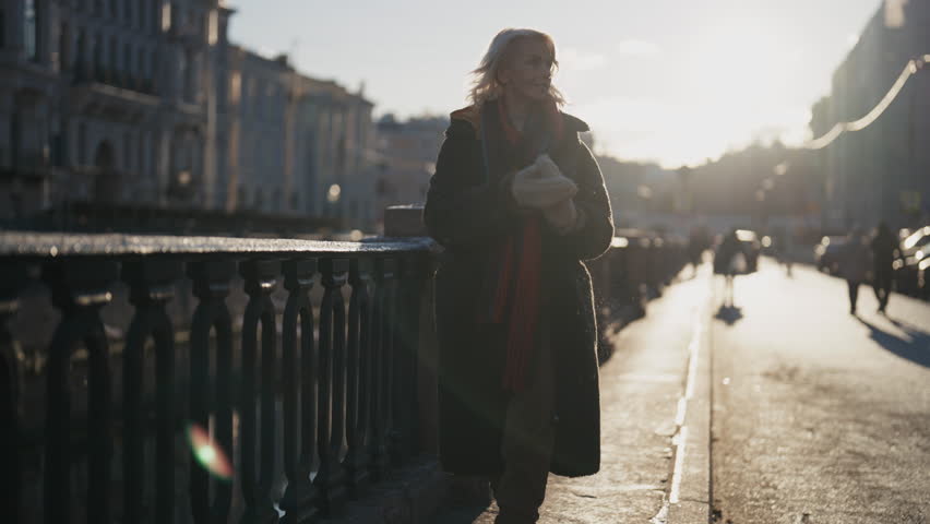 Cinematic shot with blonde woman walking in city in winter, backlit shot outdoor . Middle-aged lady enjoying good sunny weather and beauty of old city, strolling along embankment in sunny morning