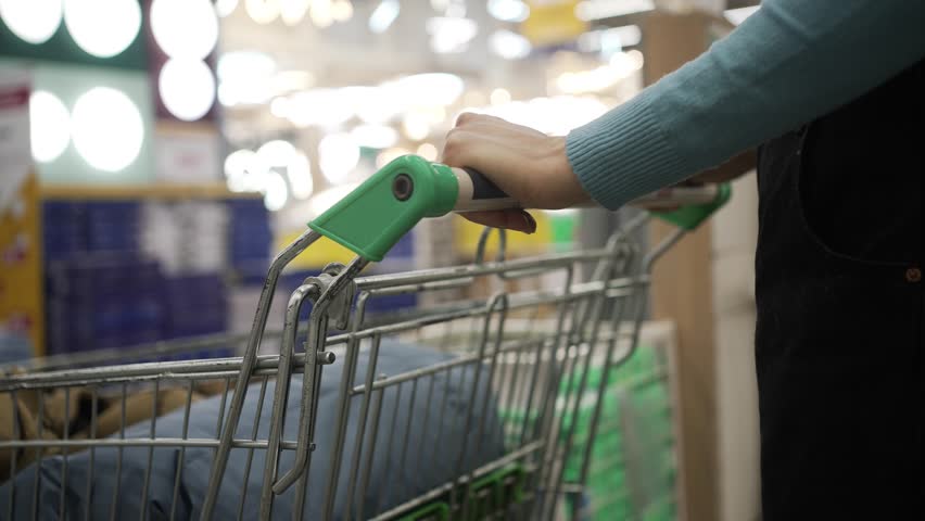 Female shopper navigating grocery store interior, pushing green-handled shopping cart through blurred retail space while selecting household items and products