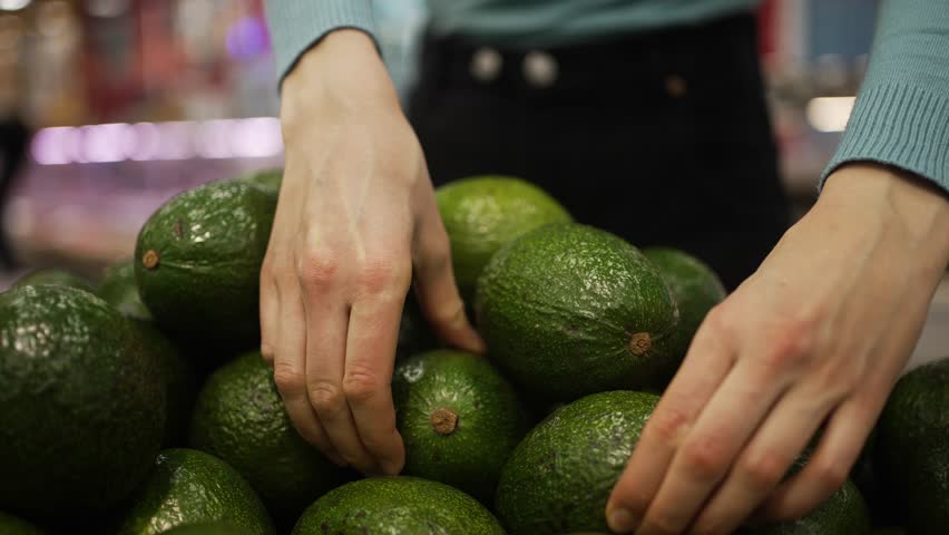 Careful customer selecting premium avocados from vibrant supermarket produce section, evaluating fruit freshness and optimal ripeness for healthy eating