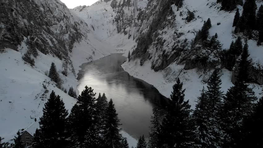 Aerial flyover above trees overlooking a frozen lake Fälensee surrounded by snow in Appenzell, Switzerland with a reflection of the winter sunset glow and mountain peaks on the dark ice