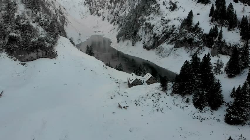 Aerial flyover over the edge of a freezing lake Fälensee surrounded by snow in Appenzell, Switzerland with a reflection of the winter sunset glow and mountain peaks on the dark ice