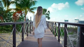 Happy blonde woman walking to sea beach, following shot lady in tropical garden. Rearview of attractive slender lady in white gown, joyful female vacationer enjoying life on ocean coastline, back view - Powered by Shutterstock - Get 15% off with code: PIKWIZARD15