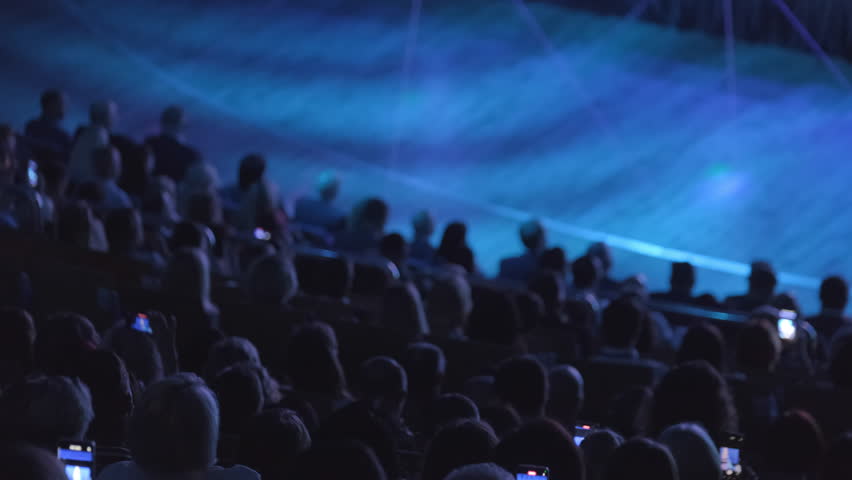 Crowd watching a stage performance from behind, with audience members capturing moments on smartphones.