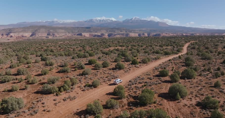 Wide aerial following vehicle across vast desert landscape on rugged remote road