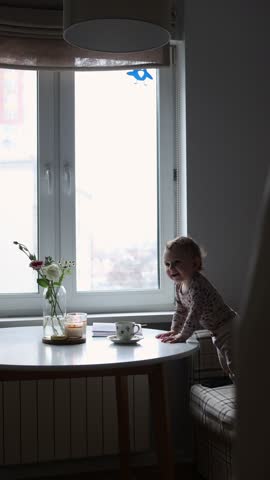 A mother in pajamas sits at the kitchen table by the window, gently takes her child in her arms, and gazes out the window at the flowers on the table. vertical video 
