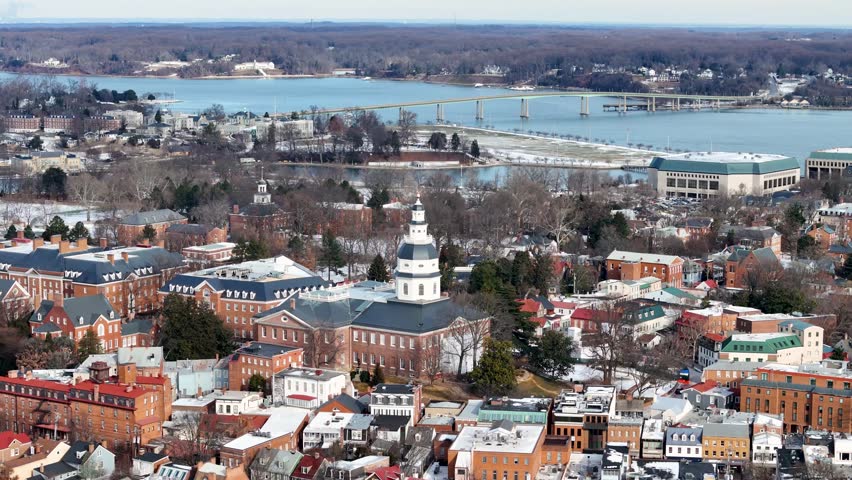 A drone glides over Annapolis, showcasing the historic State House surrounded by charming brick buildings. A bridge stretches in the distance over blue waters, with crisp winter air setting the scene