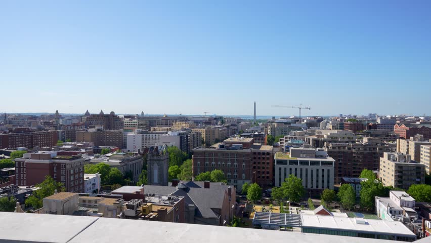 A cinematic rooftop view overlooks Washington, D.C., with the Monument standing tall in the distant background. Buildings and a crane frame the bustling cityscape under a bright, sunny sky.