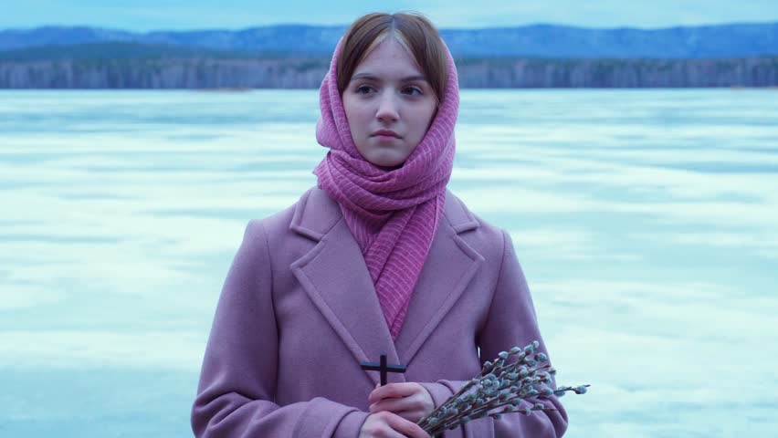 Portrait of a thoughtful young girl holding a bouquet of willow branches and an Orthodox cross. Palm Sunday, Easter. The concept of religion and human spirituality.