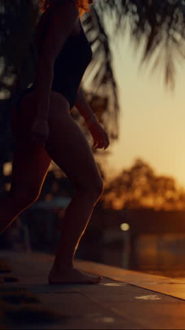 Slender woman in swimsuit jumping into outdoor swimming pool in luxury hotel, silhouette against sunset sky and sun. Vertical video with carefree female vacationer, slow motion, merry summer vacation