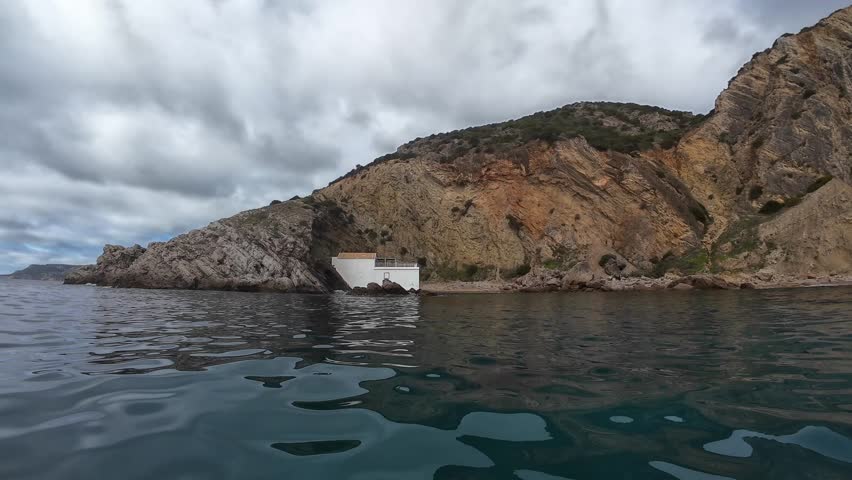 A secluded white house sits on a rugged Portuguese coastal cliff, partially embedded in the rock, with calm waters and a cloudy sky in the foreground. A hidden and unique seaside destination.