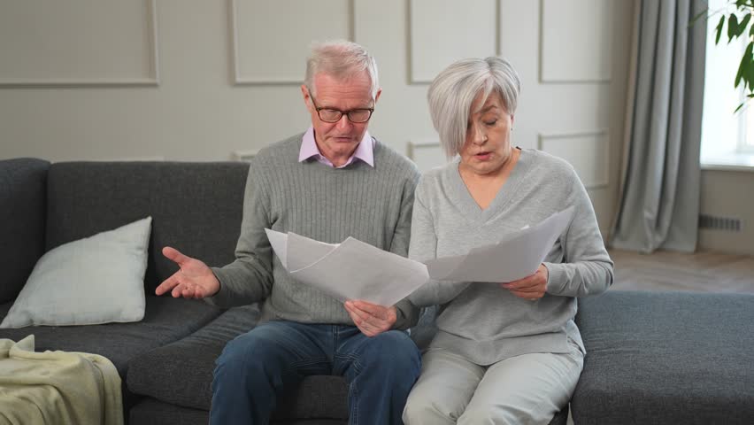 Sad tired disappointed middle aged senior couple sit with paper document. Unhappy older mature man woman reading paper bill managing bank finances calculating taxes planning loan debt pension payment