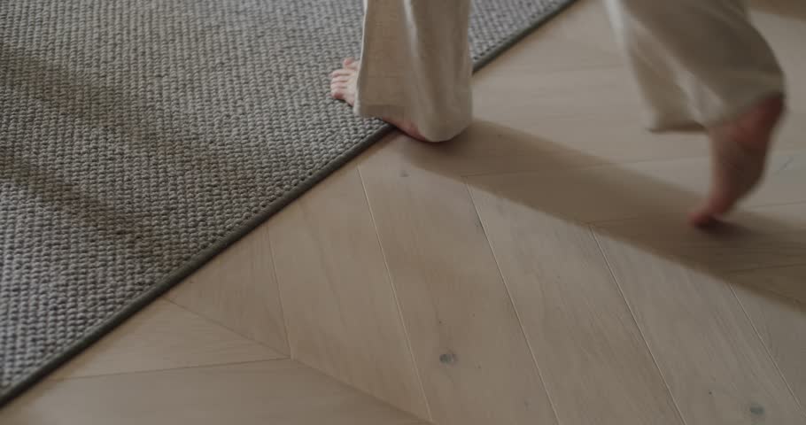 A minimalist image capturing bare feet walking on a wooden floor next to a carpet, evoking a sense of warmth and coziness in a home interior. Bare Feet Walking On Wooden Floor Next To Carpet