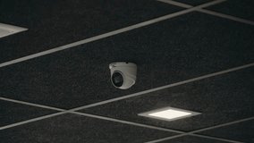 A white security camera hangs from a dark, textured ceiling. The ceiling features a grid pattern with integrated lighting. The camera is positioned centrally, suggesting surveillance and security. - Powered by Shutterstock - Get 15% off with code: PIKWIZARD15