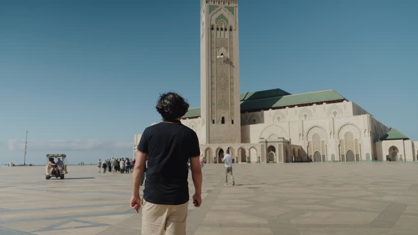 A man walks towards the Hassan II Mosque in Casablanca, Morocco. Tourists and locals enjoy the sunny day. The mosque is a beautiful example of Islamic architecture.