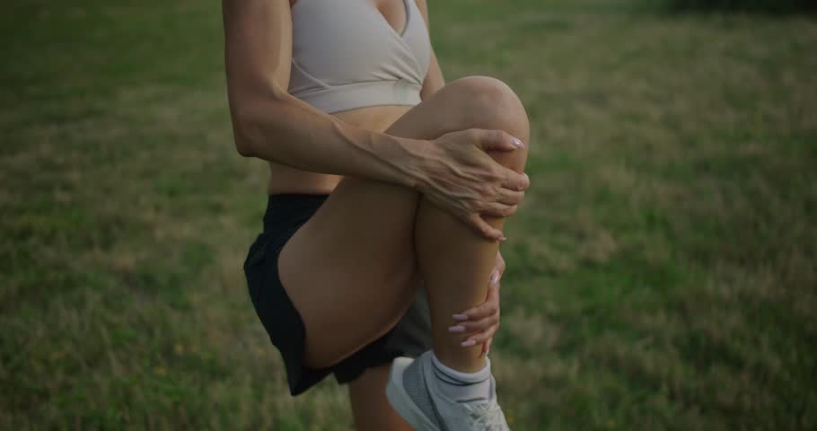 Young athletic woman warming up before workout outdoors. Sporty female in top and shorts stretch leg on stone after physical exercise in park. Wellness influencer warming up. Female stretching legs.