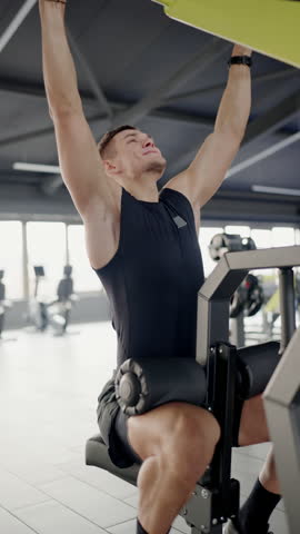 A focused young man trains intensely in a gym, displaying effort and concentration during a workout. His expression highlights the intensity of his training. Young Man Exercising Hard in Gym. Vertical