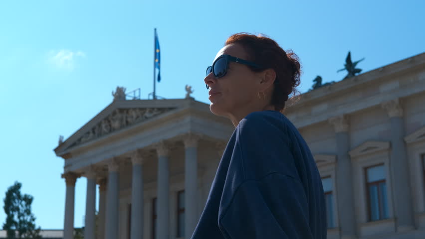 Tourist enjoying sunny day in front of austrian parliament building. Cheerful redhead woman wearing sunglasses relaxing outdoors, gazing upward with austrian parliament building under bright sky