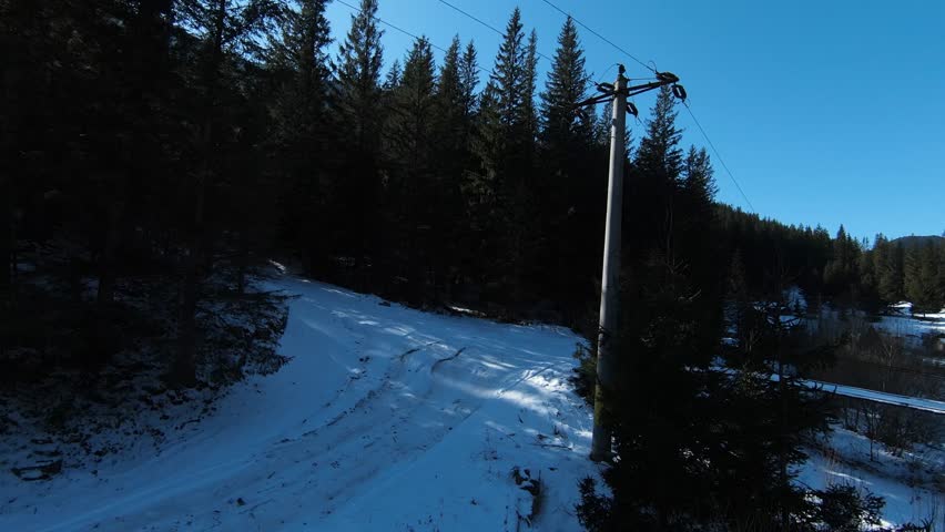 FPV drone captures a snowy path by a river, with a rustic house and forested hills under a clear blue sky, showcasing the serene beauty of a winter landscape.