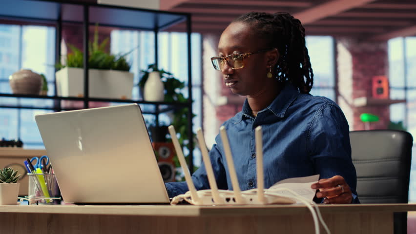 Black woman at home examines the modem and troubleshooting guide book, struggling to fix Wi Fi router and restore her internet connection for remote freelance tasks. Signal loss. Camera B.