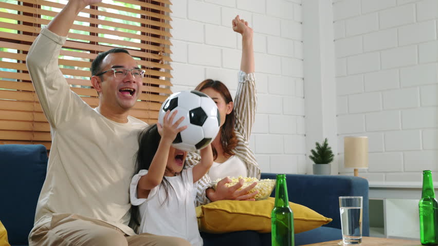 With popcorn and a ball, delighted parents and children watch a football match, shouting and celebrating. Their togetherness and smiles capture the joy of a successful goal at home.