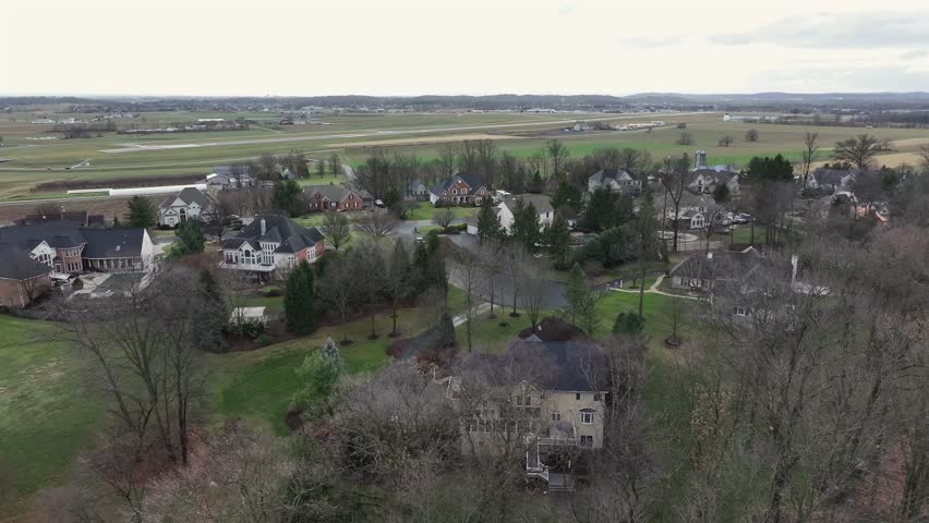 Aerial flyover luxury suburb neighborhood with villa and mansions during cloudy day. Winter day with agricultural fields in distance. Street with parking cars in america.