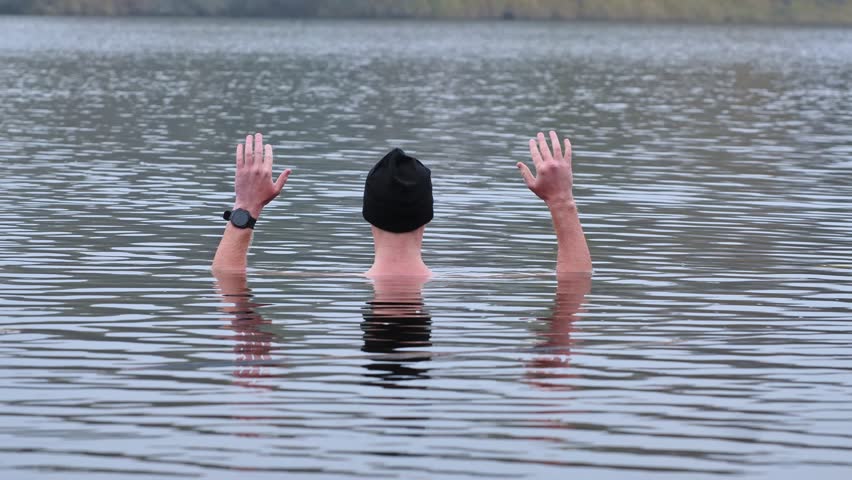 Man in freezing water, leaving only his arm with black hat visible above, 4K