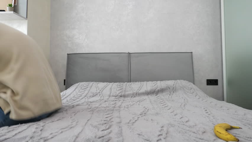 A young man eats a banana while lying on the sofa, sitting on a bed in an apartment eating a banana fruit. In slow motion.