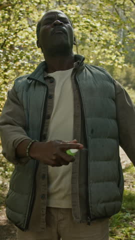 Vertical view of Black senior man spraying insect repellent on hiking clothes during outdoor activity in woods