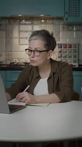 Mature Woman Writing with Left Hand While Working on Laptop from Home. Vertical Screen.