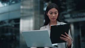 Portrait of Thai woman entering data from work papers into laptop sitting on city street. Concentrated Asian brunette businesswoman reviews corporate documents holding clipboard - Powered by Shutterstock - Get 15% off with code: PIKWIZARD15