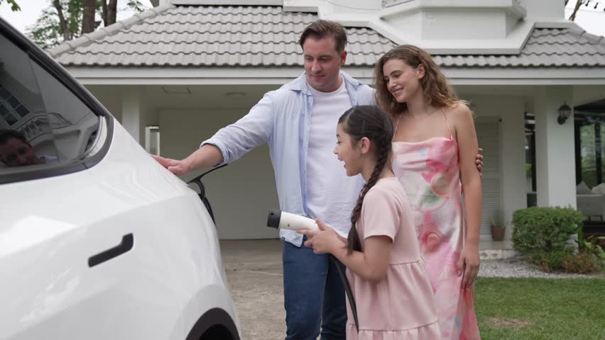 Happy little young girl learn about eco-friendly and energy sustainability as she help her family recharge electric vehicle from home EV charging station. EV car and modern family concept. Fastidious