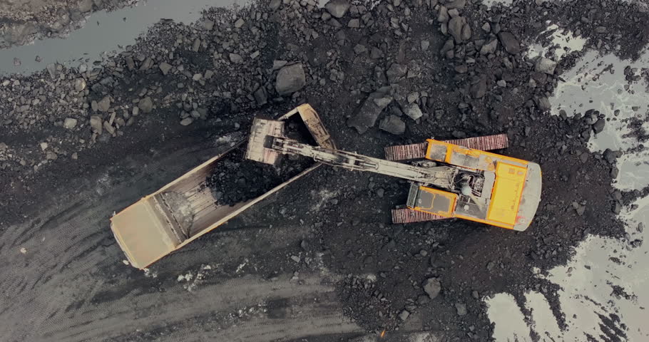 Drone view of an excavator loading coal into a truck inside an Indian coal mine. The top-angle shot captures the mining activity, highlighting heavy machinery in action amidst rugged terrain.