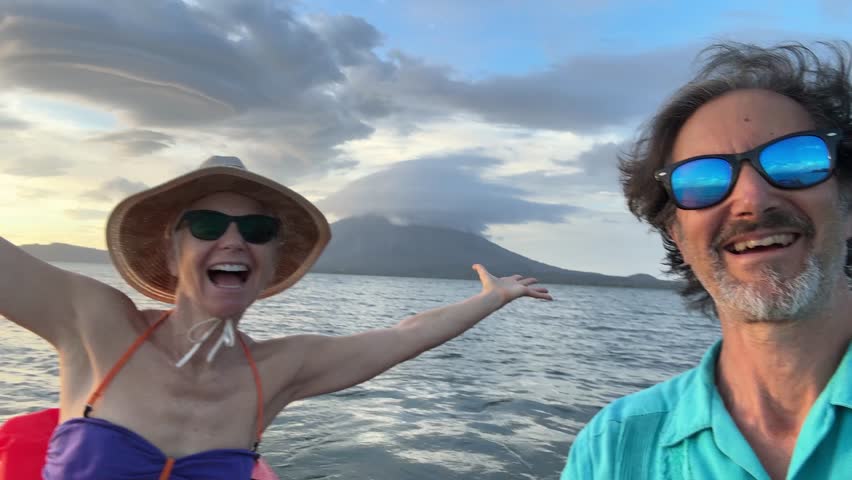 A mature man and woman share a joyful moment taking a selfie while kayaking in Lake Nicaragua during sunset. The backdrop features the stunning Conception volcano in Central America.