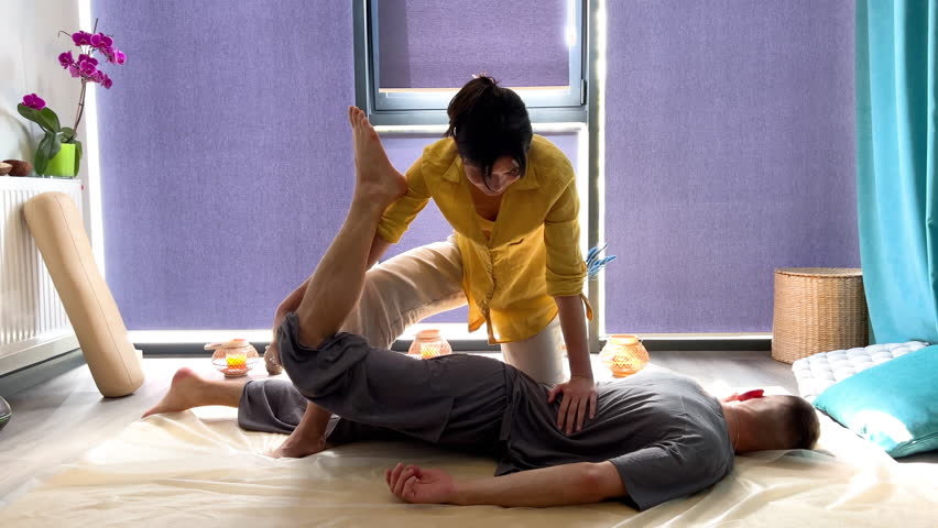 Masseuse making thai yoga massage. Woman therapist doing traditional massage treatment, stretching male patient's body. Client lying on mat, enjoying hand massage in calm atmosphere.