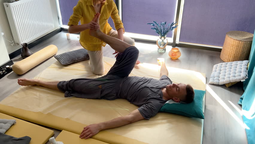 Masseuse making thai yoga massage. Woman therapist doing traditional massage treatment, stretching male patient's body. Client lying on mat, enjoying hand massage in calm atmosphere.