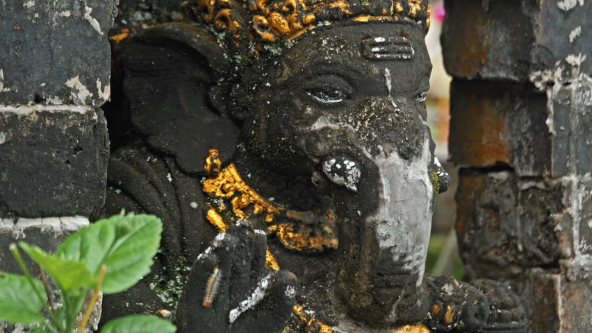 An old stone statue of Ganesha covered with moss, standing on the street with green plants