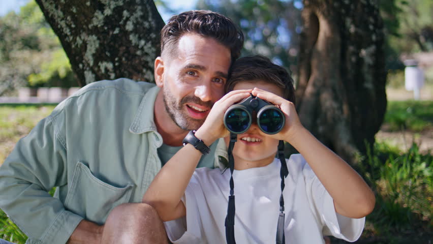 Little boy looking binoculars in forest having outdoor adventure with daddy closeup. Positive father preschool child enjoying birdwatching with smile. Man sitting under tree spending time with son