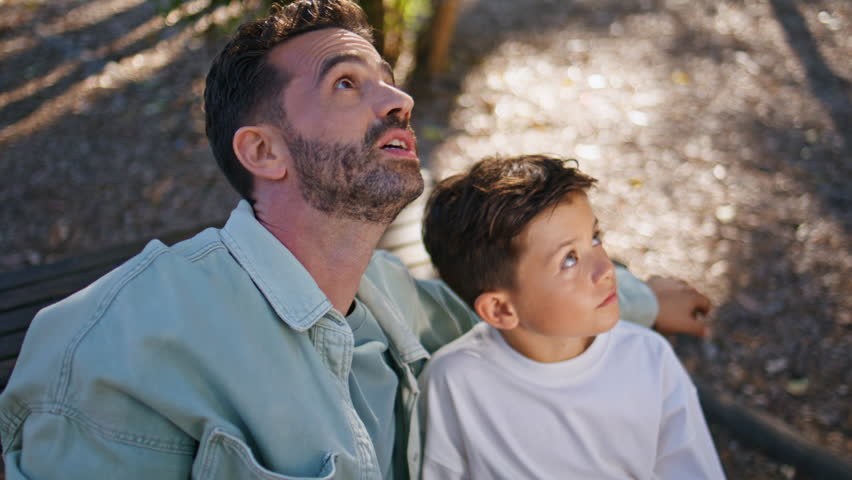 Interested father son looking distance at park weekend closeup. Caring dad pointing finger attracting attention of smiling child enjoying family moment at sunny forest. Happy man boy sitting bench