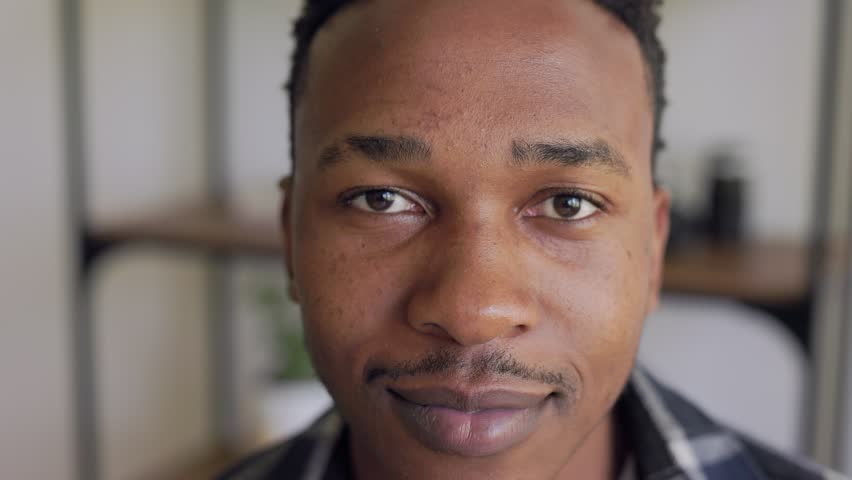 close-up portrait of a african-american man with short, curly hair and a slight smile. dark brown eyes and is wearing a checked shirt. The background is blurred, but it looks like a cozy indoor space