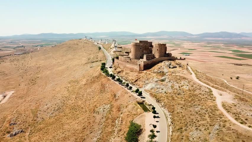 Aerial view of the medieval Consuegra Castle and its iconic windmills standing on a hilltop, overlooking the vast, dry plains of Castilla–La Mancha, Spain, a timeless symbol of Spanish heritage